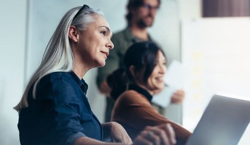 A woman with gray hair working in a collaborative setting with colleagues in the background.