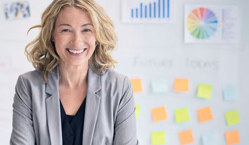 A professional woman smiling confidently while standing in an office, with colorful sticky notes in the background.